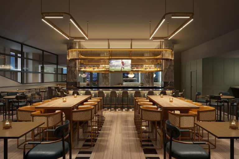 Modern hotel lounge with wooden tables, tan chairs, geometric lighting fixtures, and a gold-framed bar with TV display
