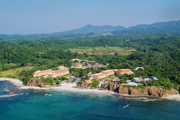 Aerial view of beachfront resort with orange-roofed buildings nestled between lush green mountains and turquoise ocean
