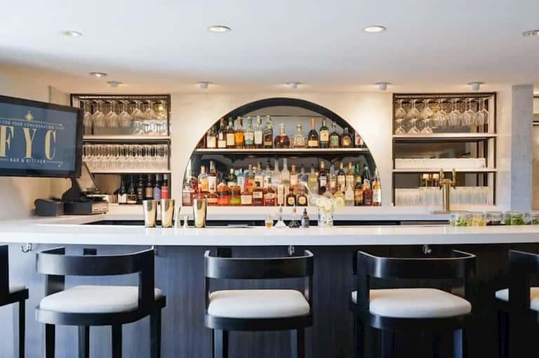 Modern bar and kitchen with white countertop, black bar stools, extensive bottle display, and glass shelving