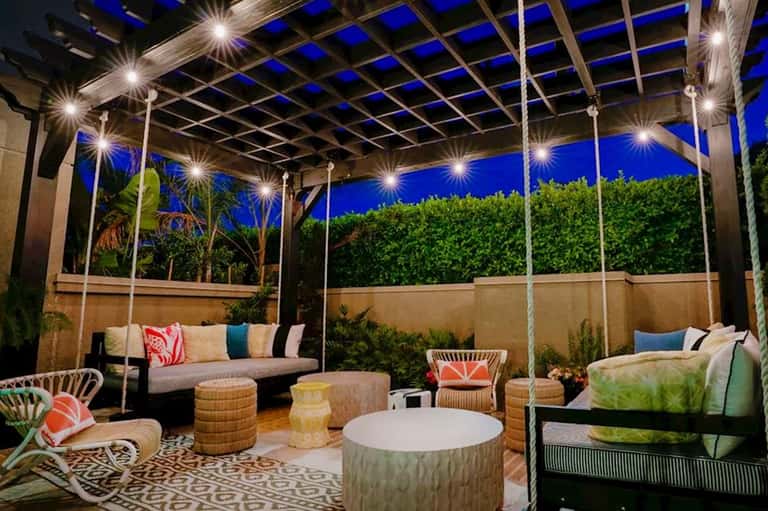 Illuminated pergola lounge area at night with colorful cushions, woven poufs, and tropical plants