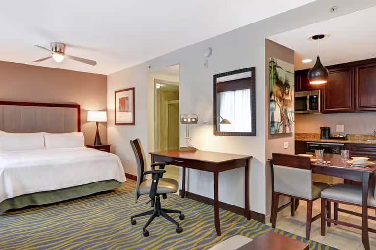 Modern hotel suite with queen bed, work desk, kitchenette, and striped area rug in warm tones