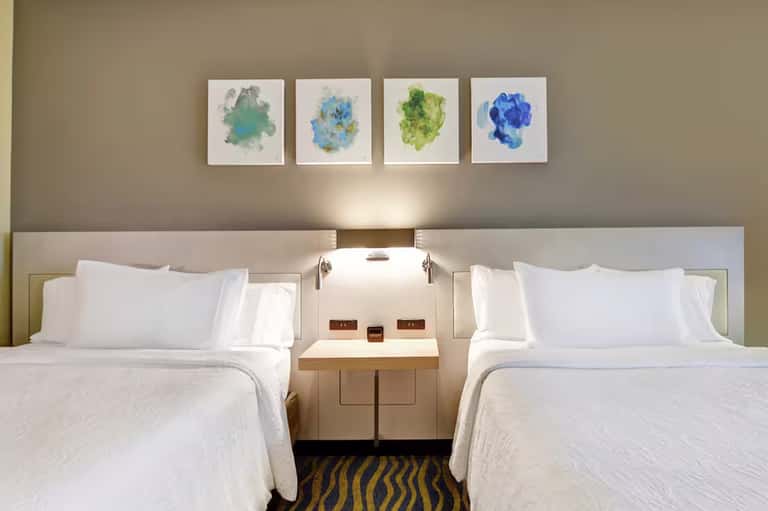 Modern hotel room with two queen beds, white linens, neutral headboard, and four colorful abstract watercolor paintings above
