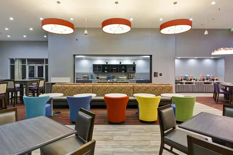 Modern hotel lobby with colorful barrel seating, orange pendant lights, and open kitchen visible in background