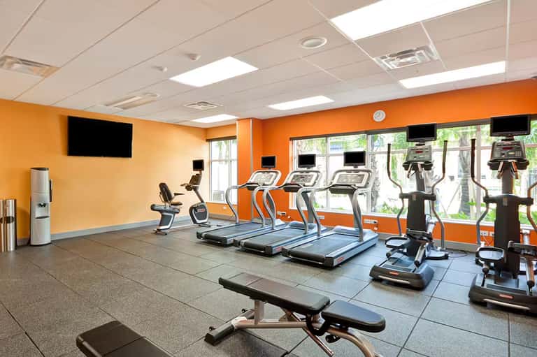 Bright orange fitness center with cardio equipment, treadmills, exercise bikes, and weight benches under white ceiling panels