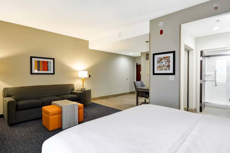 Modern hotel room with bed, sofa, orange ottoman, and ensuite bathroom