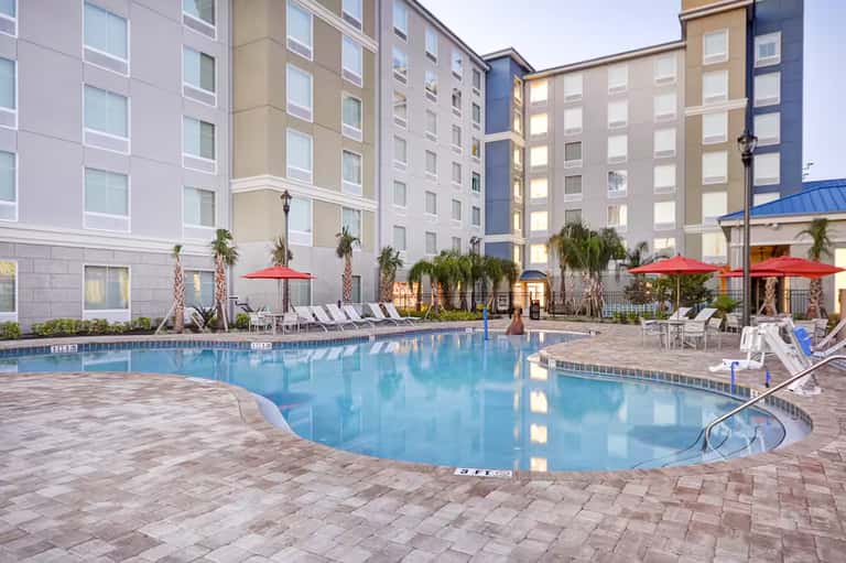 Resort courtyard with large swimming pool, lounge chairs, red umbrellas, and multi-story hotel buildings