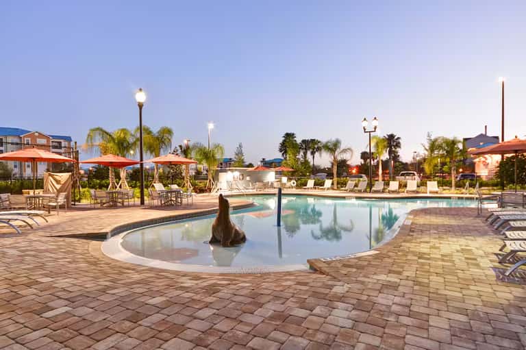 Resort pool deck at dusk with lounge chairs, umbrellas, palm trees, and lit lamp posts reflecting in calm water