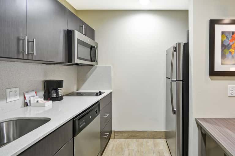 Contemporary hotel kitchenette with stainless steel appliances, dark cabinetry, and white countertops