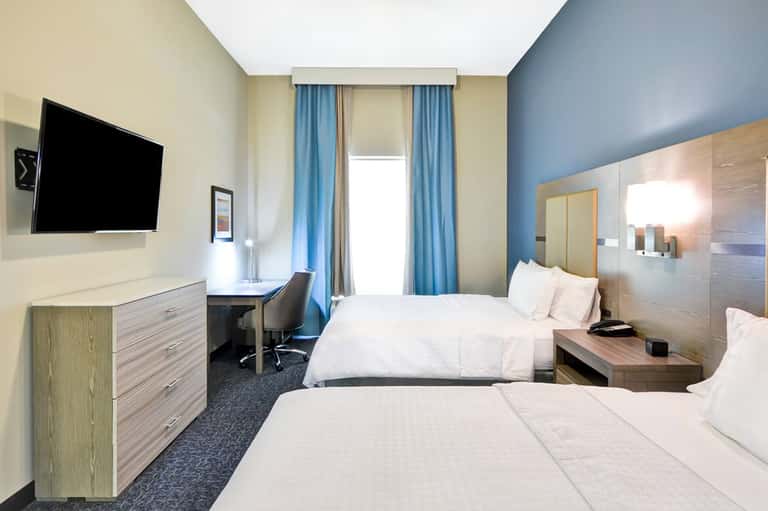 Modern hotel room with two beds, blue accent wall, flat-screen TV, and contemporary furnishings