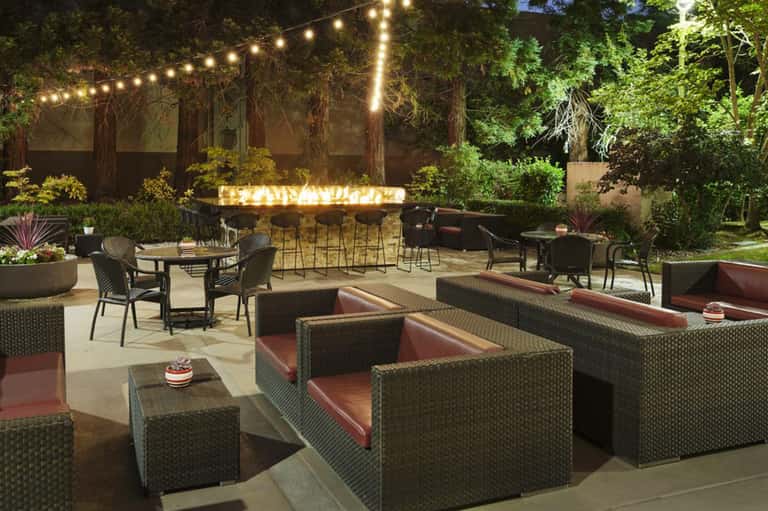 Outdoor patio at night with string lights, woven lounge seating with burgundy cushions, and illuminated bar in background