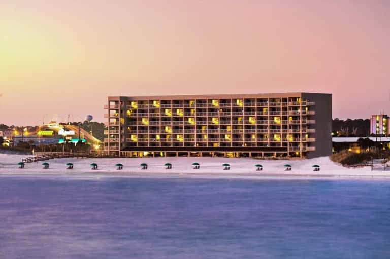 Beachfront hotel building at dusk with illuminated windows and beach umbrellas along the shore