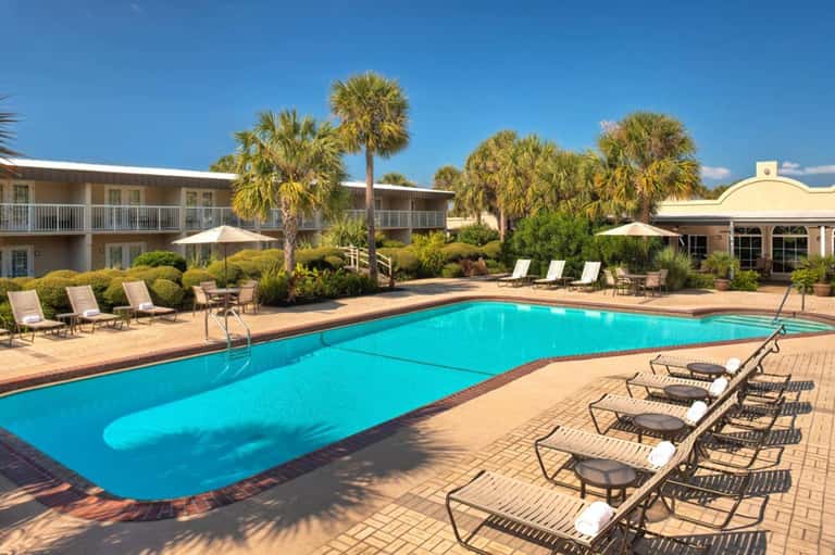Resort pool with turquoise water, lounge chairs, palm trees, and two-story buildings under clear blue sky
