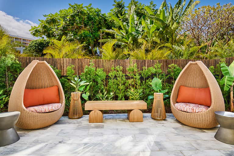 Outdoor patio with two woven egg chairs with orange cushions, wooden bench, and lush tropical plants behind wooden fence