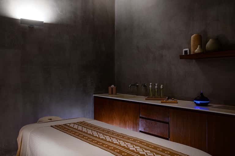 Spa treatment room with massage table, dark textured walls, wooden vanity with oils and toiletries, and ambient lighting