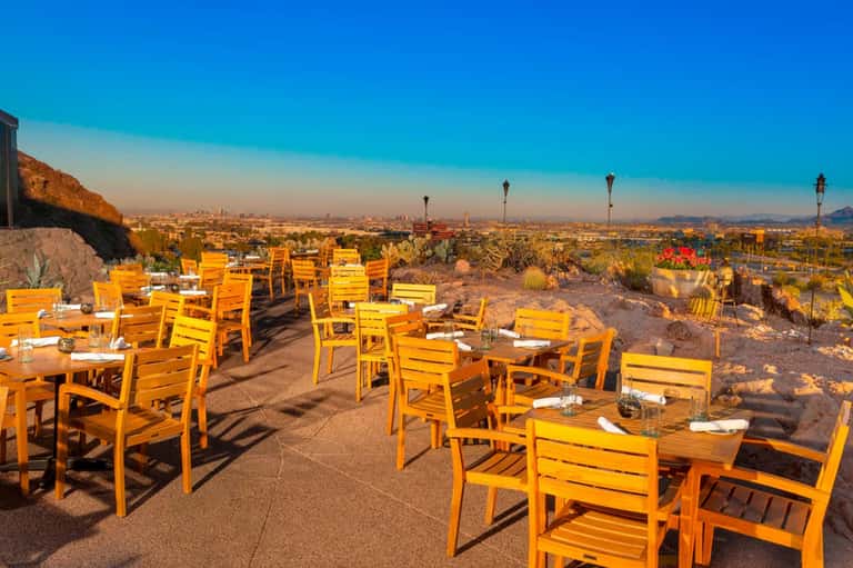 Outdoor desert dining terrace with yellow chairs overlooking sprawling cityscape at sunset