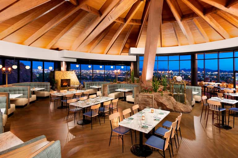 Upscale dining room with wooden cathedral ceiling, floor-to-ceiling windows overlooking city lights at dusk