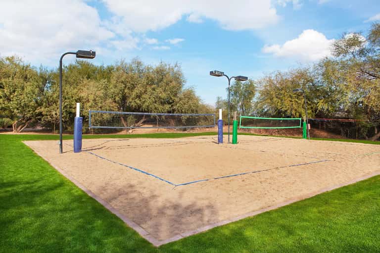 Outdoor beach volleyball court with sand, blue and green nets, surrounded by grass and palm trees