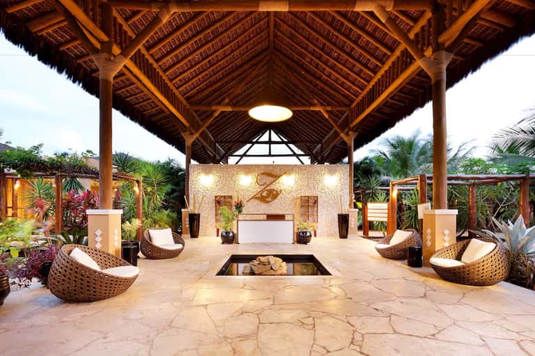 Luxury resort spa pavilion with wooden architecture, woven chairs, and stone fireplace surrounded by tropical plants