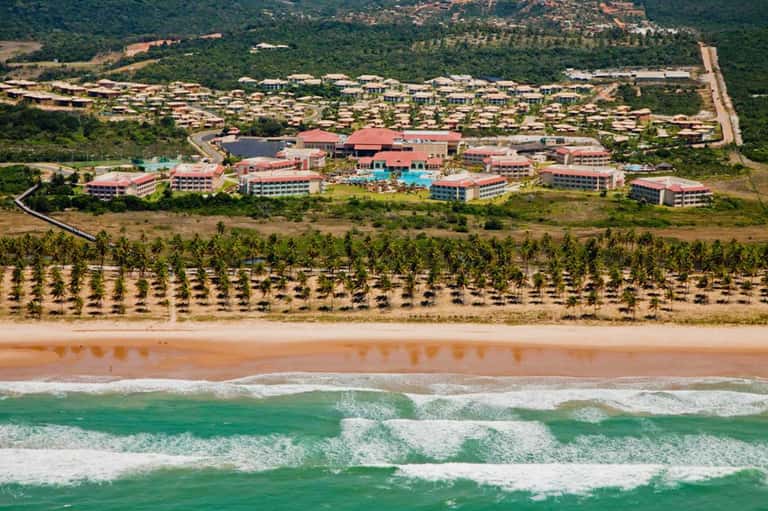 Aerial view of beachfront resort with red-roofed buildings, palm tree plantation, and ocean waves