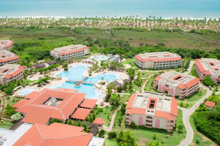 Aerial view of resort with red-roofed buildings, multiple pools, and palm trees near coastline