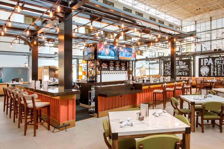 Modern brewery restaurant with orange corrugated bar, wooden chairs, high ceilings with copper pendant lights, and dining tables