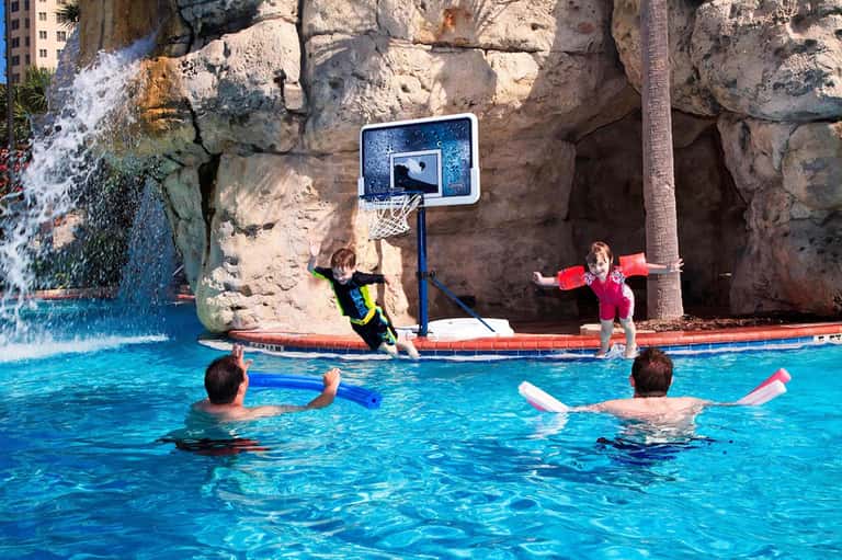 Children swimming and playing basketball in bright blue indoor pool with rock wall backdrop