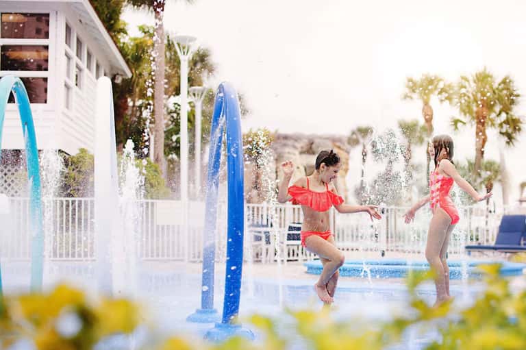 Children playing in a splash pad with water fountains near a white beachside building with palm trees