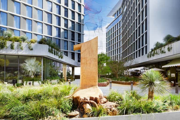 Contemporary urban plaza with modern high-rise buildings, landscaped gardens, and sculptural wooden art installation