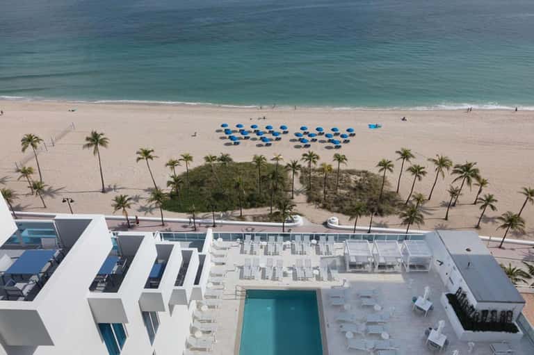 Aerial view of beachfront resort with pool, blue umbrellas, palm trees, and turquoise ocean waters