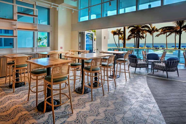 Modern beachfront dining area with floor-to-ceiling windows, wooden bar seating, and ocean views with palm trees