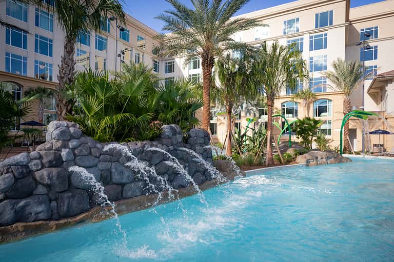 Resort pool with turquoise water, rock waterfall features, palm trees, and multi-story hotel building