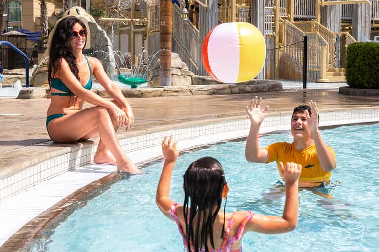 Adults and children playing with beach ball in outdoor pool area with fountains and resort buildings