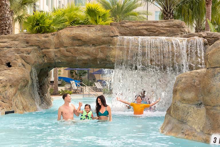 Family enjoying tropical pool with cascading waterfall and rock formations, palm trees in background
