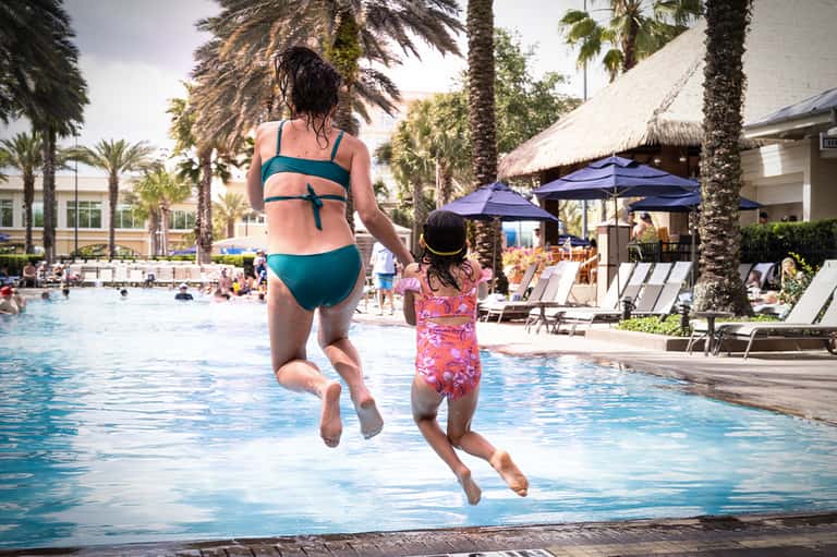 Woman and child jumping into resort pool with palm trees and blue umbrellas in background