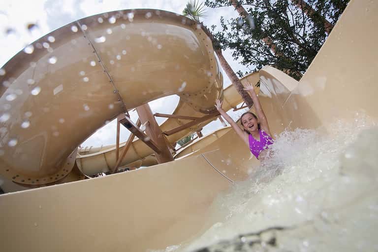 Child splashing down a large tan water slide at an outdoor water park surrounded by palm trees