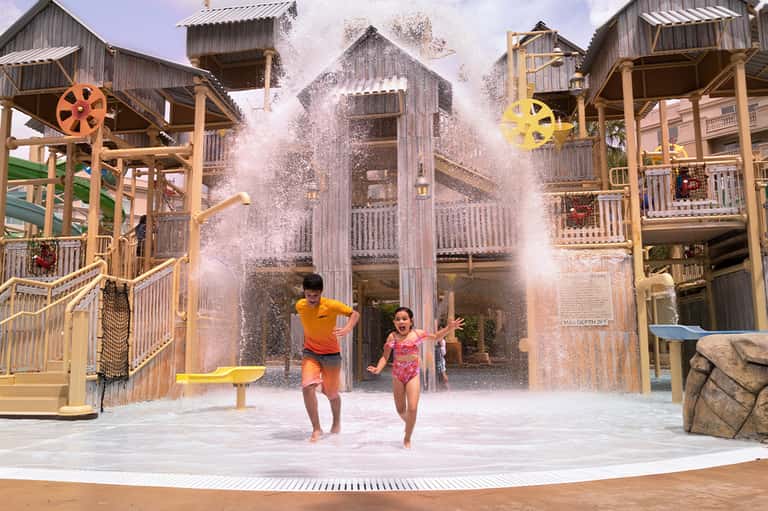 Children playing in water splash pad at resort with wooden structures and yellow wheels