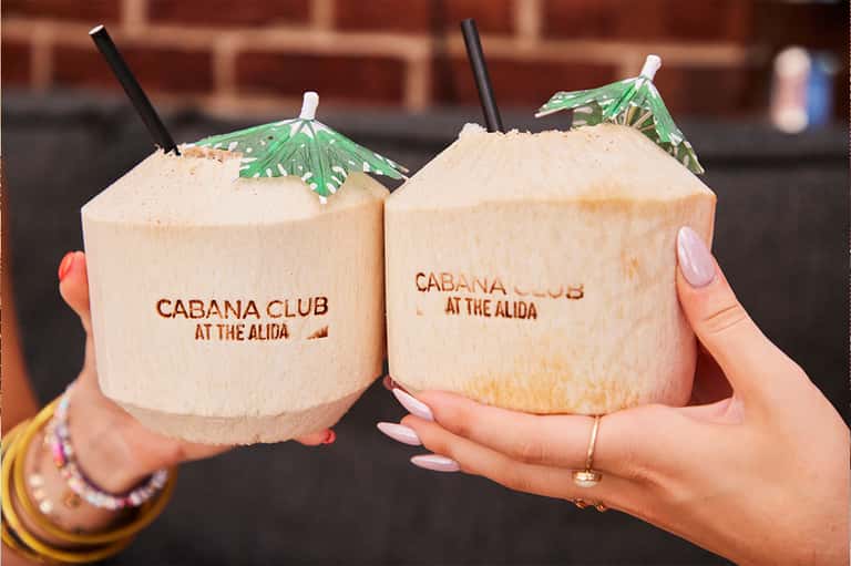 Two coconut drinks with branding from Cabana Club at The Alida, garnished with umbrellas and straws