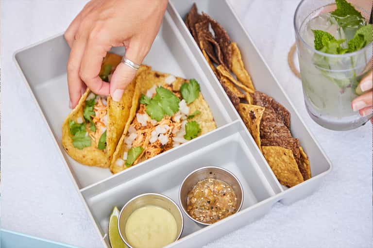 Plated tacos with shredded chicken, cilantro, and onions served with salsa and lime in a compartmentalized box