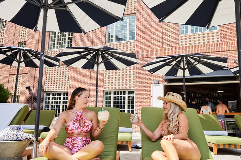 Two women relaxing on green lounge chairs under black and white umbrellas in a brick courtyard patio area.