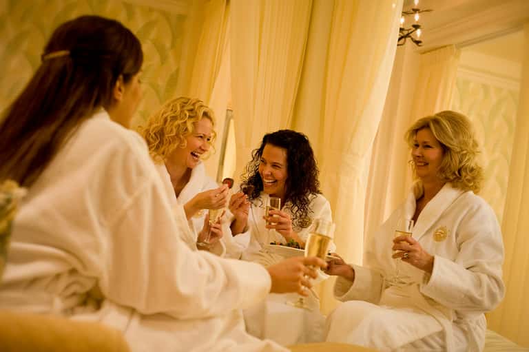Four women in white robes toasting with champagne in a luxurious spa lounge with golden curtains