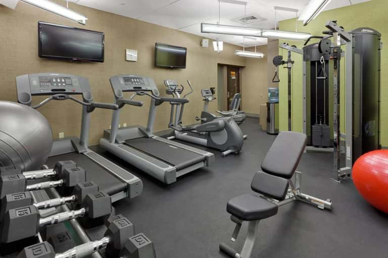 Modern fitness center with treadmills, exercise equipment, dumbbells, and TV screens on beige walls