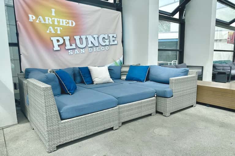 Modern rattan sectional sofa with blue cushions in hotel lobby under 'I Partied at Plunge San Diego' banner