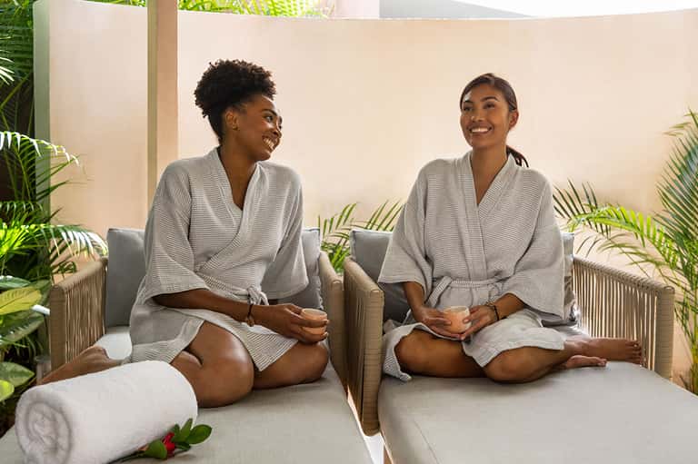 Two women in spa robes sitting on massage beds in a plant-filled wellness room