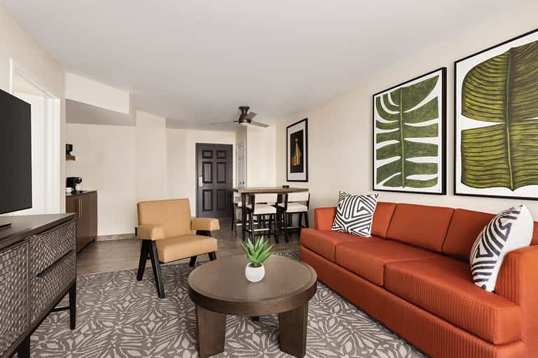 Modern hotel suite with orange sofa, tan armchair, dining area, and botanical wall art