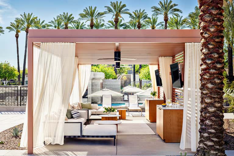 Covered outdoor lounge area with cream curtains, wooden pergola, lounge seating, and pool view surrounded by palm trees