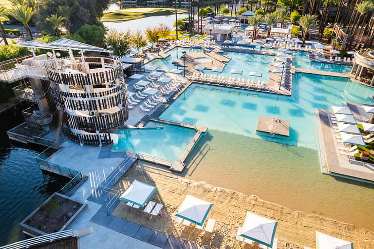 Aerial view of large resort pool complex with multiple pools, sunbathing areas, umbrellas, and tropical landscaping