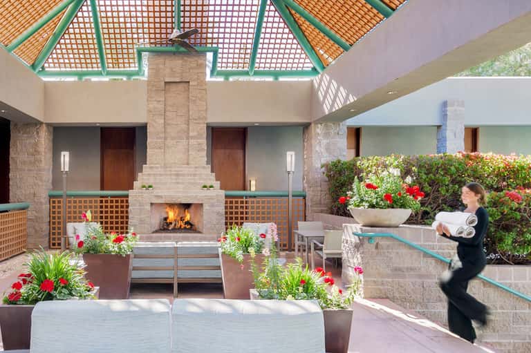 Covered courtyard with central fireplace, flowering plants, wooden doors, and latticed green ceiling