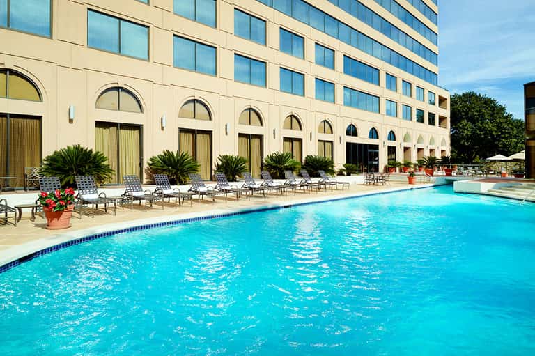 Resort outdoor pool with crystal blue water, lounge chairs, palm plants, and cream-colored building with arched windows
