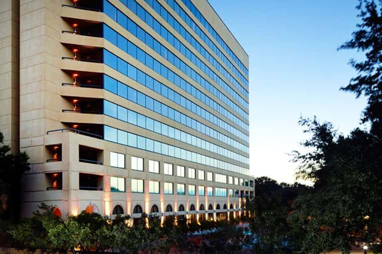 Modern multi-story hotel building with glass windows and lit balconies at dusk surrounded by landscaping