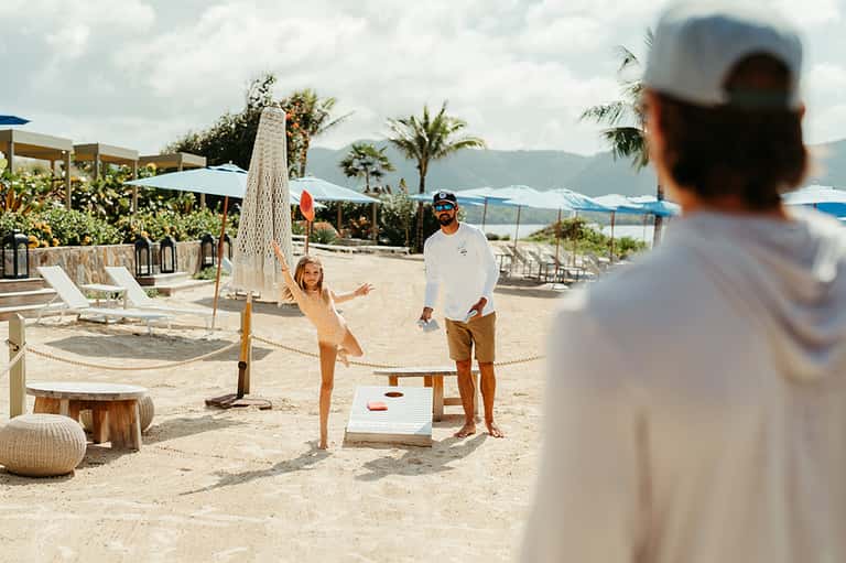Guests playing cornhole on a sandy beach with palm trees and umbrellas in background